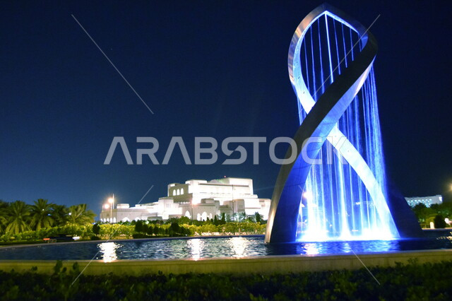 دار الأوبرا السلطانية في مسقط, معالم ثقافية وفنية سياحية مشهورة في سلطنة عمان، فن التصميم الهندسي المعماري، الحديقة الممتلئة بالأشجار والنباتات الخضراء المحيطة بالبناء، النافورة المائية المضيئة باللون الأزرق.