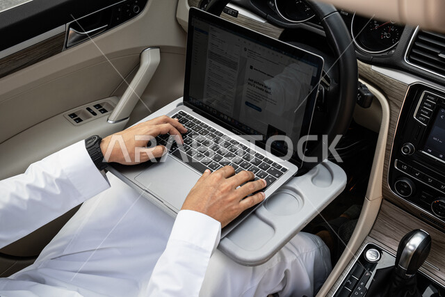 مفهوم ضغط العمل ومحاولة انجاز المهام, إدارة المشاريع عن بعد, صورة مقربة ليدين لرجل أعمال عربي خليجي سعودي يرتدي الثوب التقليدي يعمل على الحاسب المحمول في السيارة, استخدام الأجهزة والتطبيقات الحديثة  والمتطورة