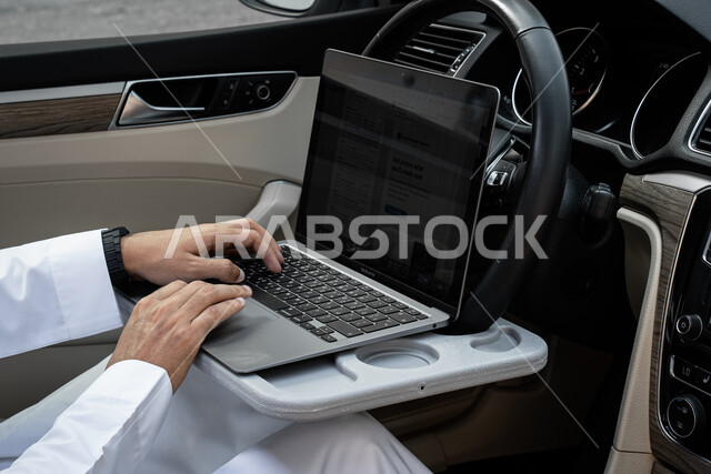 Remote project management, a close-up picture of the hands of a Saudi Arabian Gulf businessman wearing a traditional dress working on a laptop in the car, the concept of work pressure and trying to complete tasks, using modern and advanced devices and applications