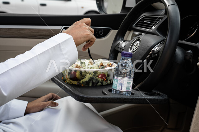 مأكولات ومقبلات باردة, سناكات صحيه خفيفه, وجبة طعام بسيطه, صورة مقربة من الجانب ليدين شاب عربي خليجي سعودي يرتدي الثوب التقليدي يتناول سلطة السيزر داخل السيارة, مزيج الخضراوات الطازجة