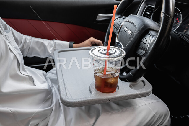 مفهوم امتلاك سيارة جديدة, الاستمتاع بالمشروبات الباردة, صورة من الجانب لشاب عربي خليجي سعودي يرتدي الثوب التقليدي يجلس داخل سيارته, وسائل النقل في المملكة العربية السعودية, كوب عصير منعش 