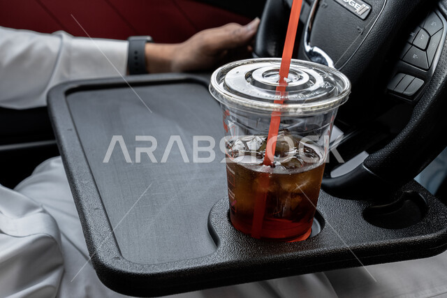 مفهوم امتلاك سيارة جديدة, صورة مقربة لكوب عصير منعش, الاستمتاع بالمشروبات الباردة, شاب عربي خليجي سعودي يرتدي الثوب التقليدي يجلس داخل سيارته, وسائل النقل في المملكة العربية السعودية