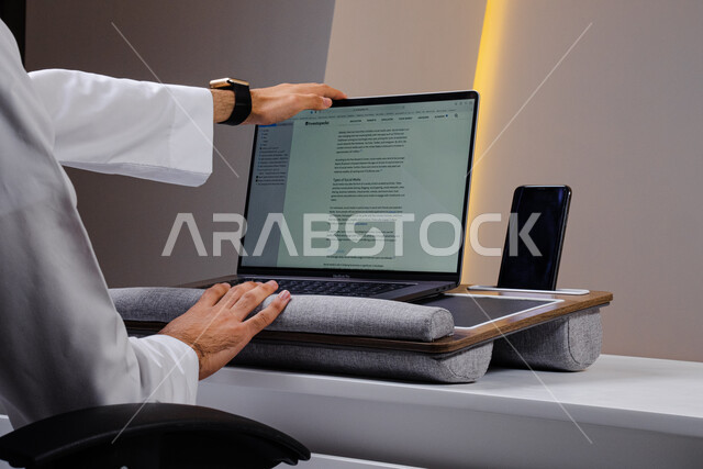 عرض الهاتف المحمول بشاشة سوداء فارغة, تنظيم جدول المهام المكتبية, مهن ووظائف إدارية, إدارة وتنفيذ المشاريع عبر أجهزة التكنولوجيا المتطورة, صورة مقربة من الخلف لشاب عربي خليجي سعودي يرتدي الثوب التقليدي يقوم باستخدام الحاسب المحمول في المكتب