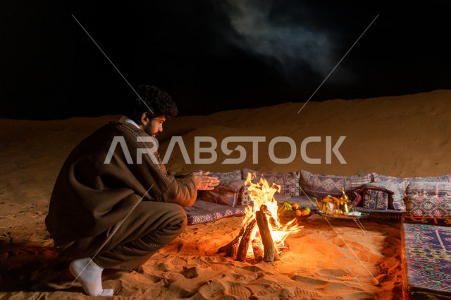 شاب سعودي يدفىء  يديه قرب شعلة الحطب , موسم الشتاء في صحاري المملكة العربية السعودية , تخييم رجل سعودي في الصحراء 