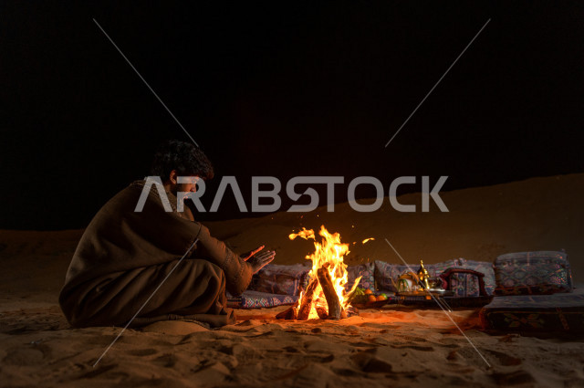 شاب سعودي يدفىء  يديه قرب شعلة الحطب , موسم الشتاء في صحاري المملكة العربية السعودية , تخييم رجل سعودي في الصحراء 
