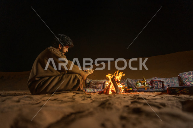 شاب سعودي يدفىء  يديه قرب شعلة الحطب , موسم الشتاء في صحاري المملكة العربية السعودية , تخييم رجل سعودي في الصحراء 