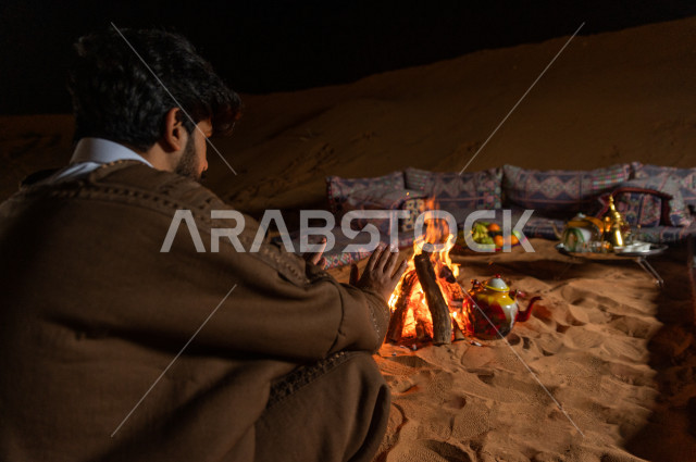 شاب سعودي يدفىء  يديه قرب شعلة الحطب , موسم الشتاء في صحاري المملكة العربية السعودية , تخييم رجل سعودي في الصحراء 