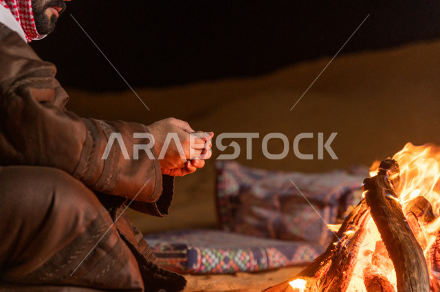 شاب سعودي يدفىء  يديه قرب شعلة الحطب , موسم الشتاء في صحاري المملكة العربية السعودية , تخييم رجل سعودي في الصحراء 