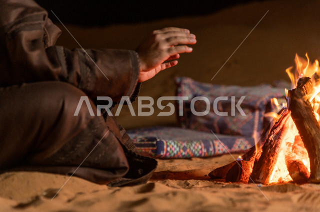 شاب سعودي يدفىء  يديه قرب شعلة الحطب , موسم الشتاء في صحاري المملكة العربية السعودية , تخييم رجل سعودي في الصحراء 