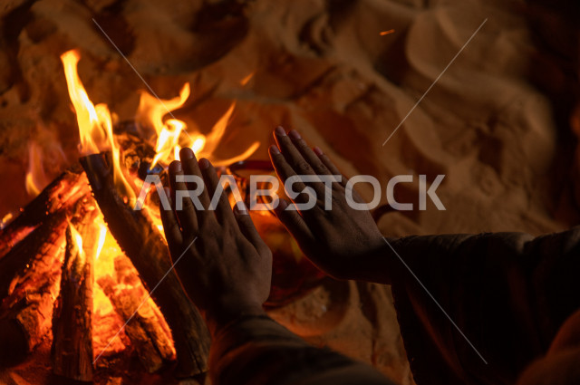 شاب سعودي يدفىء  يديه قرب شعلة الحطب , موسم الشتاء في صحاري المملكة العربية السعودية , تخييم رجل سعودي في الصحراء 