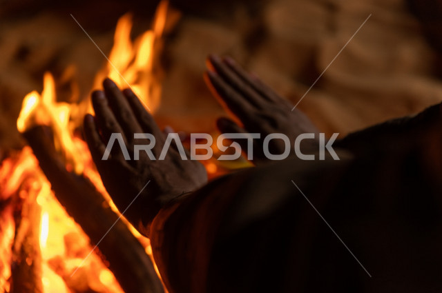 شاب سعودي يدفىء  يديه قرب شعلة الحطب , موسم الشتاء في صحاري المملكة العربية السعودية , تخييم رجل سعودي في الصحراء 
