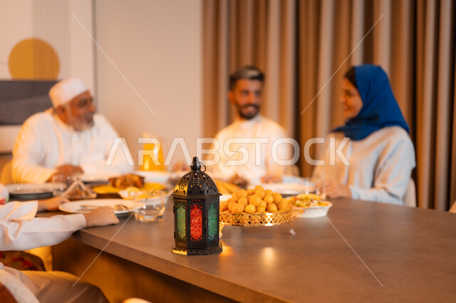 تبادل الأحاديث بين أفراد الأسرة، أجواء عائلية دينية رمضانية سعيدة، عائلة عربية خليجية سعودية مجتمعة حول مائدة الطعام، سفرة مليئة بأصناف من المأكولات الشرقية الشهية، أداء فريضة الصيام في شهر رمضان المبارك، زينة المناسبات الإسلامية