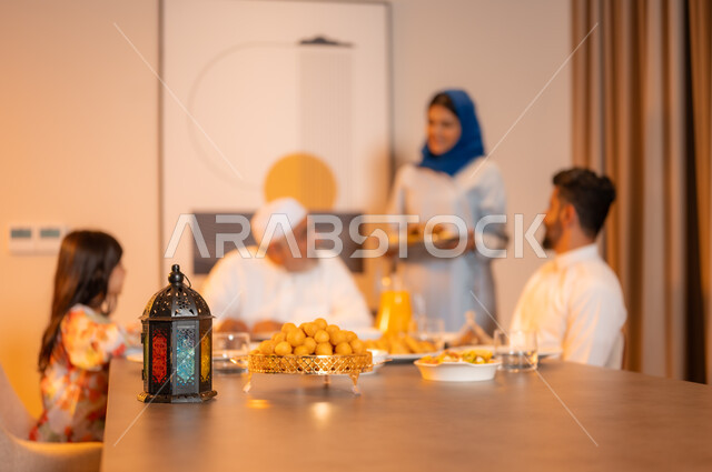 فانوس معدني مزخرف ملون على سفرة مليئة بأصناف من المأكولات الشرقية الشهية، تبادل الأحاديث بين أفراد الأسرة، أجواء عائلية دينية رمضانية سعيدة، عائلة عربية خليجية سعودية مجتمعة حول مائدة الطعام، أداء فريضة الصيام في شهر رمضان المبارك، زينة المناسبات الإسلامية