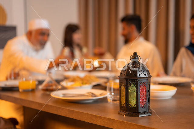 فانوس معدني مزخرف ملون على سفرة مليئة بأصناف من المأكولات الشرقية الشهية، تبادل الأحاديث بين أفراد الأسرة، أجواء عائلية دينية رمضانية سعيدة، عائلة عربية خليجية سعودية مجتمعة حول مائدة الطعام، أداء فريضة الصيام في شهر رمضان المبارك، زينة المناسبات الإسلامية