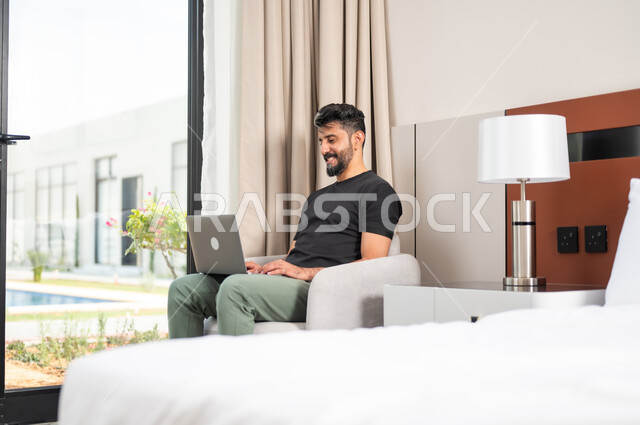Making video calls, using modern devices and technologies, a smiling Saudi Gulf Arab young man sitting on a comfortable sofa using a laptop, completing work tasks remotely, chatting and communicating with friends, browsing the Internet and social networking sites