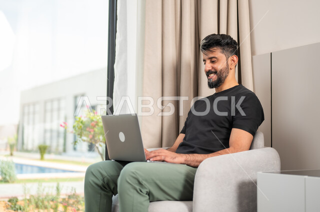 اجراء مكالمات الفيديو، استخدام الأجهزة والتقنيات الحديثة، شاب عربي خليجي سعودي مبتسم يجلس على كنبة مريحة يستخدم جهاز الحاسوب المحمول، انجاز مهام العمل عن بعد، الدردشة والتواصل مع الاصدقاء، تصفح الانترنت ومواقع التواصل الاجتماعي