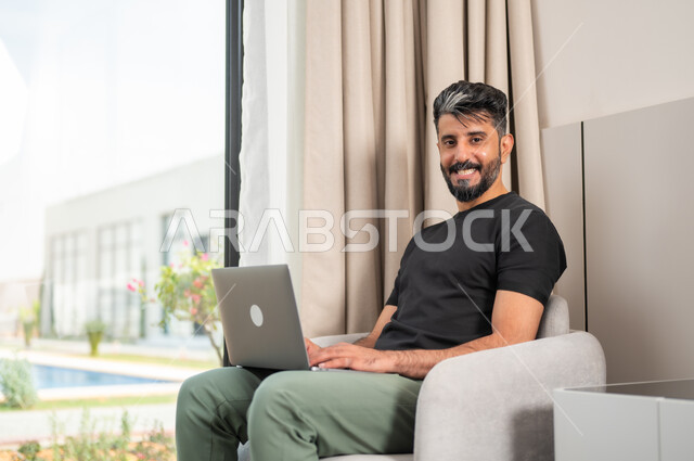 Completing work tasks remotely, using modern devices and technologies, making video calls, a smiling Saudi Gulf Arab young man looking at the camera, sitting on a comfortable sofa, using a laptop, chatting and communicating with friends, browsing the Internet and social networking sites