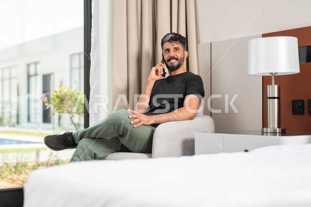 Making phone calls, using modern devices and technologies, a smiling Saudi Gulf Arab man sitting on a comfortable sofa holding a mobile phone in his hand, chatting and communicating with friends with gestures of happiness and joy, browsing the Internet and social networking sites