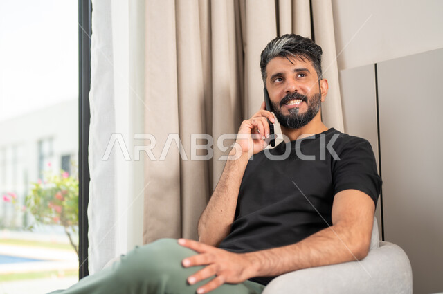 إجراء المكالمات الهاتفية، استخدام الأجهزة والتقنيات الحديثة، رجل عربي خليجي سعودي مبتسم يجلس على كنبة مريحة يمسك بيده الهاتف المحمول، الدردشة والتواصل مع الاصدقاء بإيماءات السعادة والفرح، تصفح الانترنت ومواقع التواصل الاجتماعي
