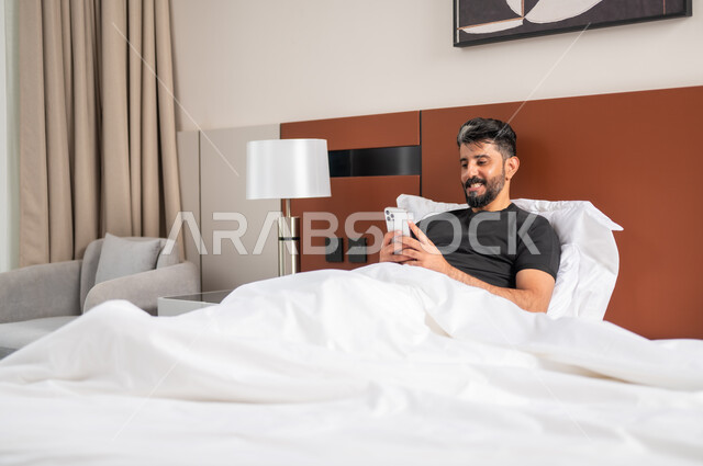 استخدام الأجهزة والتقنيات الحديثة، رجل عربي خليجي سعودي مبتسم يجلس على السرير يمسك بيده الهاتف المحمول، الدردشة والتواصل مع الاصدقاء بإيماءات السعادة والفرح، تصفح الانترنت ومواقع التواصل الاجتماعي