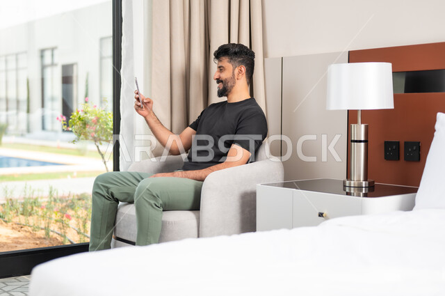 الدردشة والتواصل مع الاصدقاء بإيماءات السعادة والفرح، استخدام الأجهزة والتقنيات الحديثة، رجل عربي خليجي سعودي مبتسم يجلس على كنبة مريحة يمسك بيده الهاتف المحمول، تصفح الانترنت ومواقع التواصل الاجتماعي