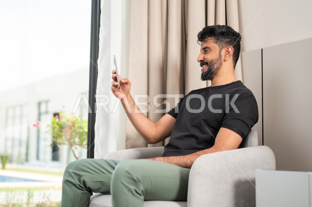 اجراء مكالمة فيديو، الدردشة والتواصل مع الاصدقاء بإيماءات السعادة والفرح، استخدام الأجهزة والتقنيات الحديثة، رجل عربي خليجي سعودي مبتسم يجلس على كنبة مريحة يمسك بيده الهاتف المحمول، تصفح الانترنت ومواقع التواصل الاجتماعي