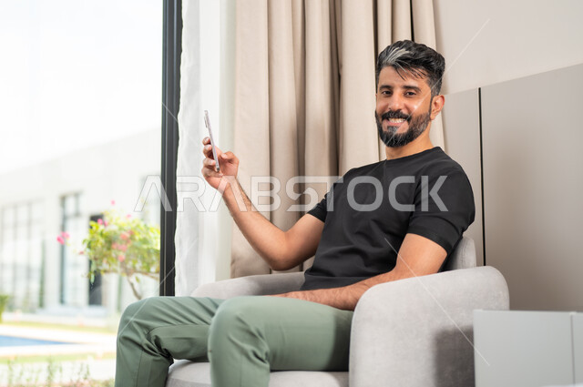 تصفح الانترنت ومواقع التواصل الاجتماعي، استخدام الأجهزة والتقنيات الحديثة، رجل عربي خليجي سعودي مبتسم يجلس على كنبة مريحة ينظر الى الكاميرا يمسك بيده الهاتف المحمول، الدردشة والتواصل مع الاصدقاء بإيماءات السعادة والفرح