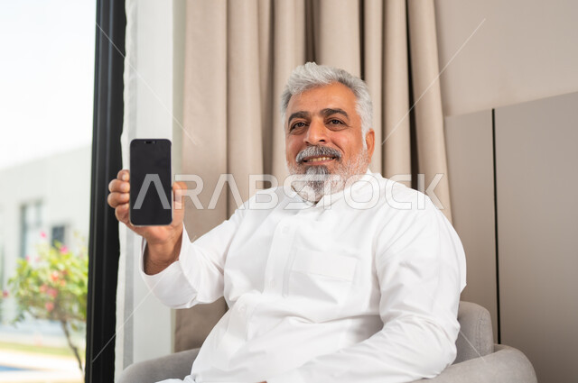 استخدام الأجهزة والتقنيات الحديثة، صورة مقربة لرجل كبير السن عربي خليجي سعودي مبتسم يرتدي الثوب التقليدي يجلس على كنبة مريحة يقوم بعرض الهاتف المحمول بشاشة سوداء فارغة، برامج وتطبيقات الجوال، تصفح مواقع التواصل الاجتماعي