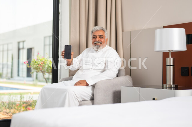 Using modern devices and technologies, an elderly smiling Saudi Arabian Gulf man wearing a traditional thobe sits on a comfortable sofa, viewing a mobile phone with a blank black screen, mobile programs and applications, browsing social networking sites