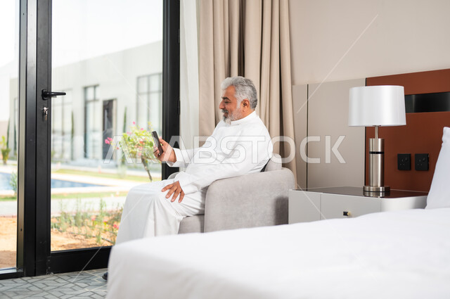 الدردشة والتواصل مع الاصدقاء بإيماءات السعادة والفرح، استخدام الأجهزة والتقنيات الحديثة، صورة مقربة لرجل كبير السن عربي خليجي سعودي مبتسم يجلس على كنبة مريحة يمسك بيده الهاتف المحمول، تصفح الانترنت ومواقع التواصل الاجتماعي