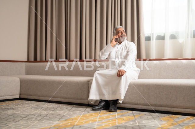التواصل والدردشة مع الاصدقاء بإيماءات السعادة والفرح، إجراء المكالمات الهاتفية، استخدام الأجهزة والتقنيات الحديثة، رجل كبير السن عربي خليجي سعودي مبتسم يجلس على كنبة مريحة يمسك بيده الهاتف المحمول، تصفح الانترنت ومواقع التواصل الاجتماعي