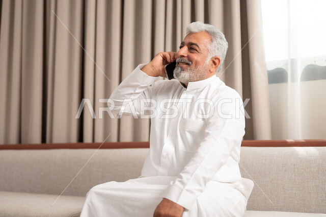 مكالمات صوتية عبر الجوال، استخدام الأجهزة والتقنيات الحديثة، صورة مقربة لرجل كبير السن عربي خليجي سعودي مبتسم يجلس على كنبة مريحة يمسك بيده الهاتف المحمول، الدردشة والتواصل مع الاصدقاء بإيماءات السعادة والفرح، تصفح الانترنت ومواقع التواصل الاجتماعي