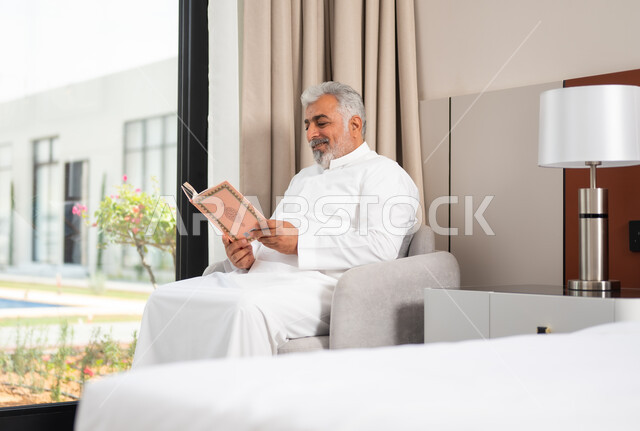 العبادة والتقرب الى الله، رجل كبير السن عربي خليجي سعودي مبتسم يرتدي الثوب التقليدي يجلس على كنبة مريحة يقوم بقراءة القرآن الكريم بإيماءات السعادة والسرور، تحفيظ وتلاوة كتاب الله، مفهوم التدبر والخشوع