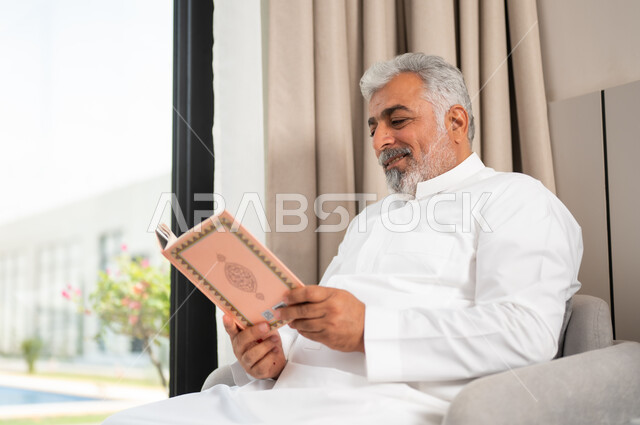 تحفيظ وتلاوة كتاب الله، العبادة والتقرب الى الله، صورة مقربة لرجل كبير السن عربي خليجي سعودي مبتسم يرتدي الثوب التقليدي يجلس على كنبة مريحة يقوم بقراءة القرآن الكريم بإيماءات السعادة والسرور، مفهوم التدبر والخشوع