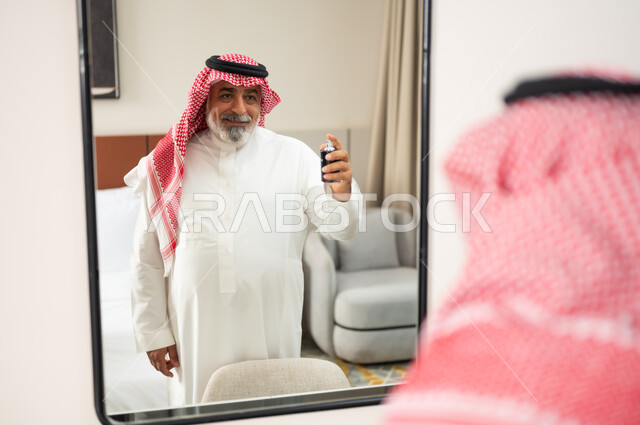 التطيب بالروائح الفاخرة، الاهتمام بالأناقة والمظهر الخارجي، صورة مقربة من الخلف لرجل عربي سعودي خليجي يرتدي الشماغ والثوب التقليدي يقف بجانب المرآة يمسك بيده العبوة الزجاجية يقوم برش العطر، مفهوم المناسبات والأعياد والاحتفالات