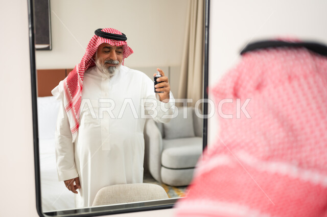 التطيب بالروائح الفاخرة، الاهتمام بالأناقة والمظهر الخارجي، صورة مقربة من الخلف لرجل عربي سعودي خليجي يرتدي الشماغ والثوب التقليدي يقف بجانب المرآة يمسك بيده العبوة الزجاجية يقوم برش العطر، مفهوم المناسبات والأعياد والاحتفالات