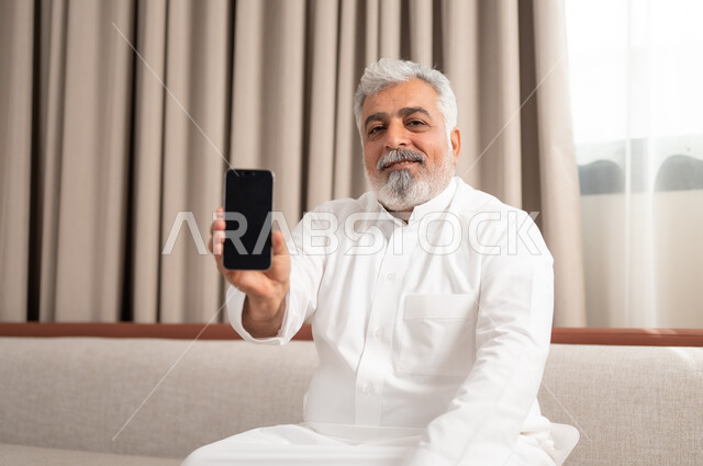 The concept of the development of technology and modern techniques, a close-up image of an elderly Saudi Arabian Gulf man, smiling, wearing a traditional dress, sitting on a comfortable sofa, displaying a mobile phone with a blank black screen, mobile programs and applications, browsing social networking sites