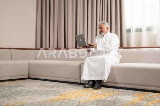 Making a video call, completing remote work tasks, using modern devices and technologies, a smiling old Saudi Gulf Arab man sitting on a comfortable sofa, using a laptop, chatting and communicating with friends, browsing the Internet and social networking sites