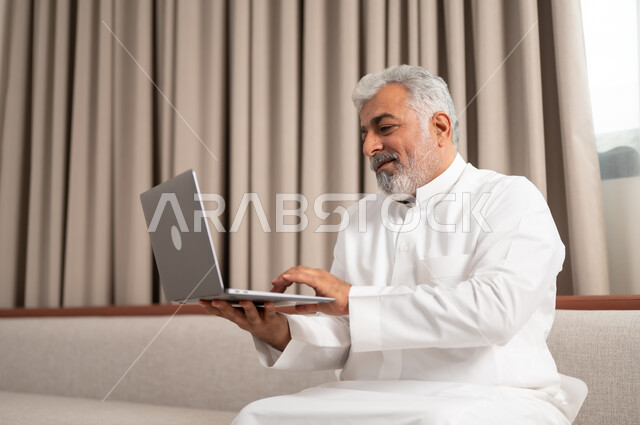 اجراء مكالمة فيديو، انجاز مهام العمل عن بعد، استخدام الأجهزة والتقنيات الحديثة، صورة مقربة لرجل شايب عربي خليجي سعودي مبتسم يجلس على كنبة مريحة يستخدم جهاز الحاسوب المحمول، الدردشة والتواصل مع الاصدقاء، تصفح الانترنت ومواقع التواصل الاجتماعي