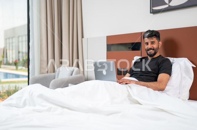 Completing work and tasks remotely, relaxing and lying down in free time, spending a comfortable and enjoyable time on vacation, using advanced modern technical devices, a smiling Saudi Gulf Arab young man lying on the bed in the bedroom holding the laptop.