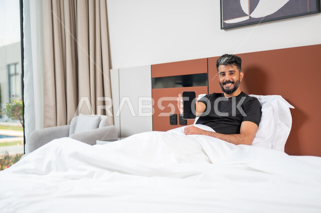 عرض هاتف محمول بشاشة سوداء فارغه، الاسترخاء والاستلقاء في اوقات الفراغ، قضاء وقت مريح وممتع في العطلة، استخدام الأجهزة التقنية الحديثة المتطورة، شاب عربي خليجي سعودي يستلقي على السرير في غرفة النوم يحمل بيده موبايل