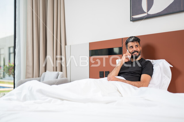 Making phone calls, using modern devices and technologies, a smiling Saudi Gulf Arab man lying on the bed, answering calls via mobile phone, chatting and communicating with friends with gestures of happiness and joy.