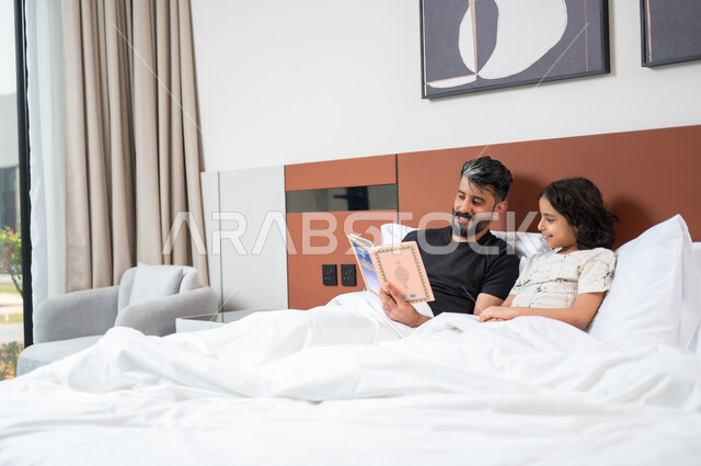 تعليم الاولاد على التثقيف والمطالعة، قضاء وقت ممتع مع الأبناء، أب عربي خليجي سعودي يجلس على السرير بجانب ابنه يقومان بقراءة القصص والحكايات قبل النوم، شغف المطالعة للكتب والروايات، القيام بالانشطة المسلية في المنزل، 