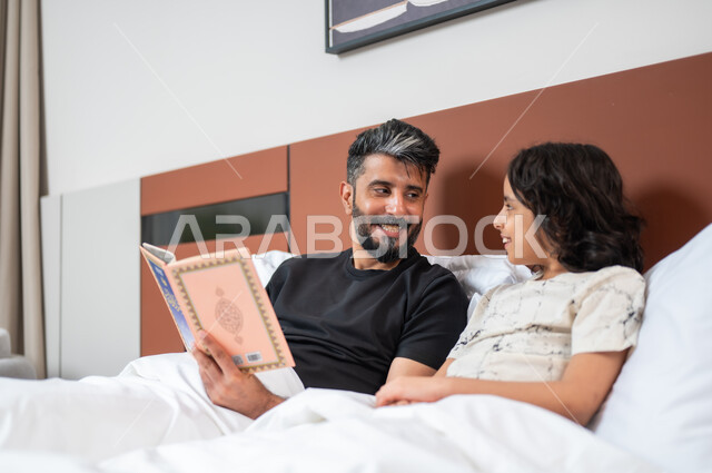 شغف المطالعة للكتب والروايات، القيام بالانشطة المسلية في المنزل، تعليم الاولاد على التثقيف والمطالعة، قضاء وقت ممتع مع الأبناء، أب عربي خليجي سعودي يجلس على السرير بجانب ابنه يقومان بقراءة القصص والحكايات قبل النوم