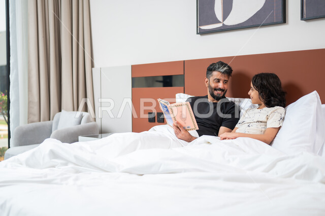 تعليم الاولاد على التثقيف والمطالعة، قضاء وقت ممتع مع الأبناء، أب عربي خليجي سعودي يجلس على السرير بجانب ابنه يقومان بقراءة القصص والحكايات قبل النوم، شغف المطالعة للكتب والروايات، القيام بالانشطة المسلية في المنزل، 