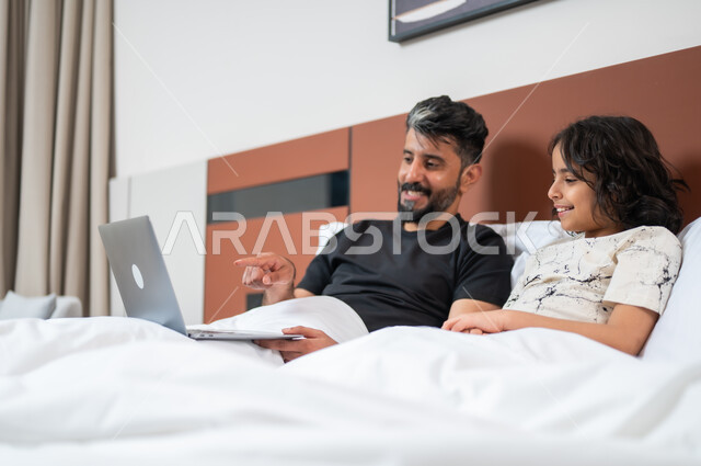 القيام بالانشطة المسلية في المنزل، مشاهدة الافلام والبرامج الترفيهيه، استخدام التكنولوجيا والأجهزة التقنية الحديثة، قضاء وقت ممتع مع الأبناء، أب عربي خليجي سعودي يجلس على السرير بجانب ابنه يقومان باجراء مكالمة فيديو عبر الحاسوب المحمول