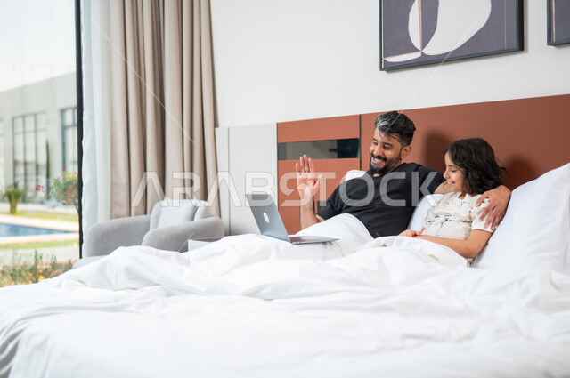Using technology and modern technical devices, spending quality time with the children, a Saudi Arabian Gulf father sitting on the bed next to his son, making a video call via the laptop, watching movies and entertainment programs, doing entertaining activities at home.