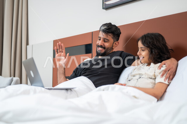 مشاهدة الافلام والبرامج الترفيهيه، القيام بالانشطة المسلية في المنزل، استخدام التكنولوجيا والأجهزة التقنية الحديثة، قضاء وقت ممتع مع الأبناء، أب عربي خليجي سعودي يجلس على السرير بجانب ابنه يقومان باجراء مكالمة فيديو عبر الحاسوب المحمول