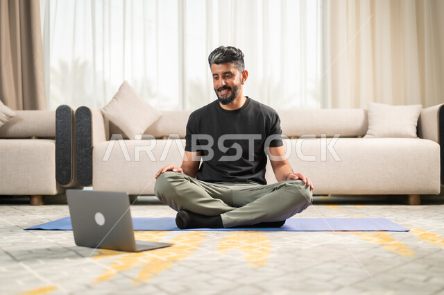 متابعة تمارين اليوغا عبر الحاسوب المحمول، استخدام الأجهزة التقنية الحديثة، الاستمتاع في قضاء الوقت في المنزل، الاسترخاء والشعور بالراحة،التدرب على الهدوء وتمالك النفس، المحافظة على اللياقة والصحة البدنية، رجل عربي خليجي سعودي يقوم بممارسة اليوغا في غرفة المعيشة