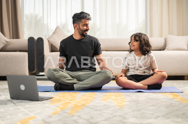 الاسترخاء والشعور بالراحة، تعليم الاولاد الهدوء وتمالك النفس، المحافظة على اللياقة والصحة البدنية، الاستمتاع في قضاء الوقت مع الأبناء، أب عربي خليجي سعودي يجلس أمام الحاسوب المحمول يقوم بممارسة اليوغا مع ابنه في غرفة المعيشة